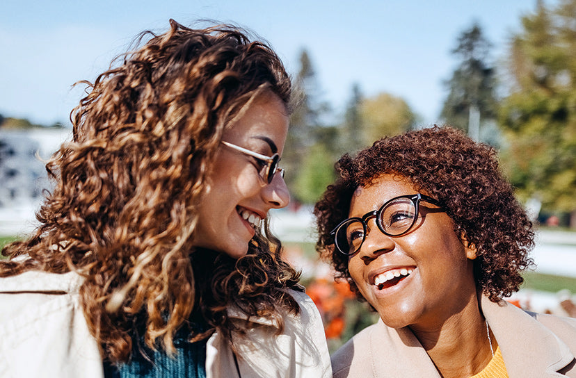 Two women smiling and laughing outdoors, both wearing round eyeglasses. One has long wavy brown hair and wears a light coat over a blue top, while the other has short curly brown hair and wears a beige top.