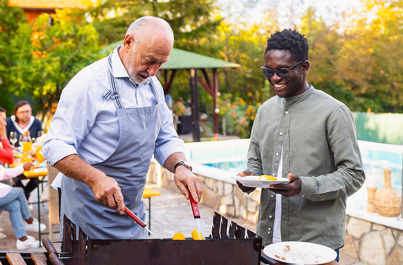 A lifestyle scene at an outdoor poolside barbecue featuring two men. One man in a light blue shirt and gray apron is grilling on a black barbecue grill, while the other man in a sage green shirt and sunglasses holds a white plate with food, smiling.
