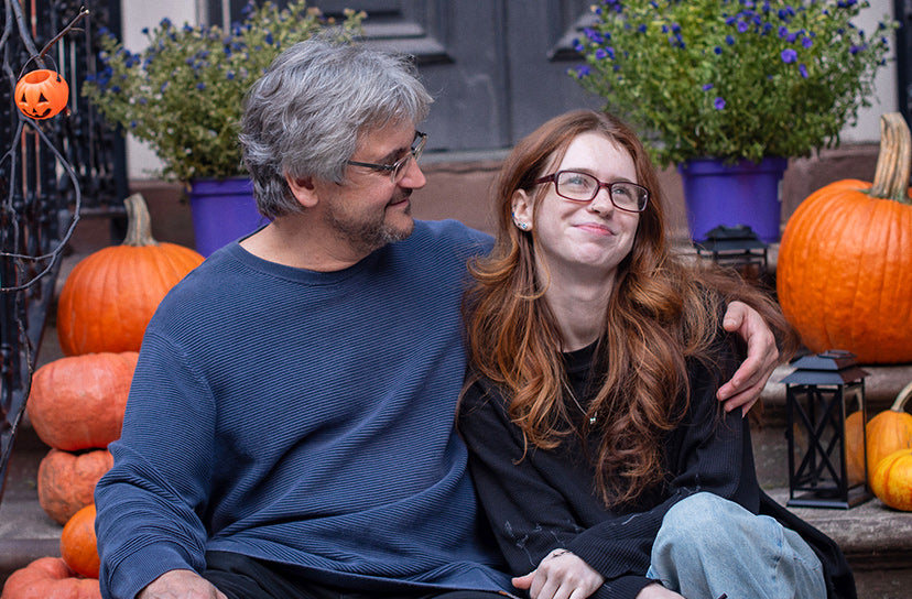 Man and woman wearing rectangular eyeglasses seated together on porch steps surrounded by pumpkins and fall flowers.
